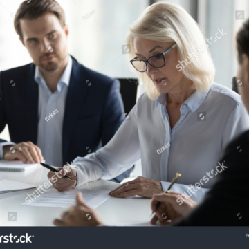 stock-photo-businessmen-sitting-at-desk-headed-by-middle-aged-serious-concentrated-female-in-eyeglasses-1249021717